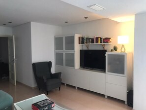 Een woonkamer met een zwarte fauteuil, een flatscreen televisie, een boekenkast en een lamp.