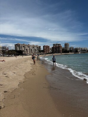 A beach with a sandy shore, a few people walking, and a cityscape with buildings in the background.