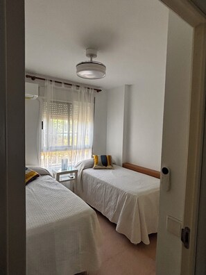 A room with two beds, a window with sheer curtains, a bedside table with a lamp, and a ceiling fan.