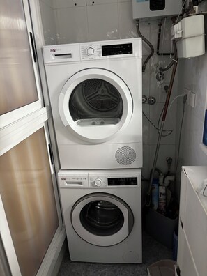 Eine übereinander gestellte Waschmaschine und Trockner in einem Waschraum, mit sichtbarer Sanitärinstallation und Warmwasserbereiter.