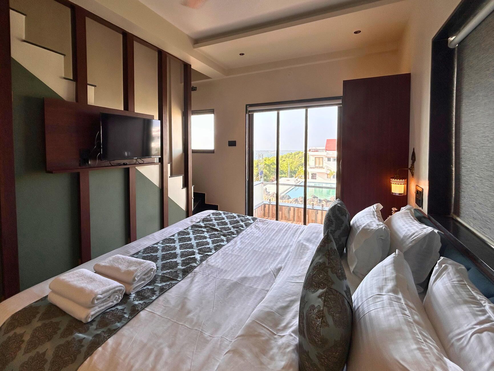 A hotel room with a bed, a television, a balcony with a pool view, and a cityscape in the background.