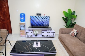 A living room with a flat-screen TV on a stand, a coffee table with a remote, a sofa with a decorative pillow, and a water dispenser. - Affordable 1 Bedroom Apartment in Utawala Nairobi Near JKIA (Nairobi)