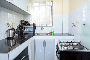 A kitchen with a gas stove, a sink, and a window with sheer curtains. - Affordable 1 Bedroom Apartment in Utawala Nairobi Near JKIA (Nairobi)