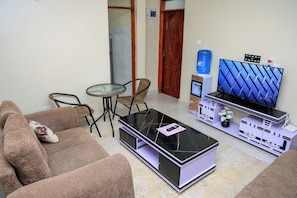 A living room with a sofa, a coffee table, a TV stand with a flat-screen TV, and a water dispenser. - Affordable 1 Bedroom Apartment in Utawala Nairobi Near JKIA (Nairobi)