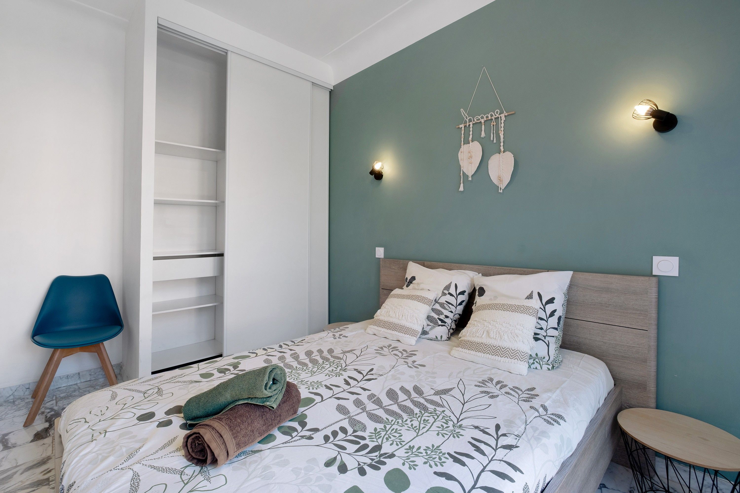 Un dormitorio con cama, una silla, una mesita de noche y una lámpara de pared.