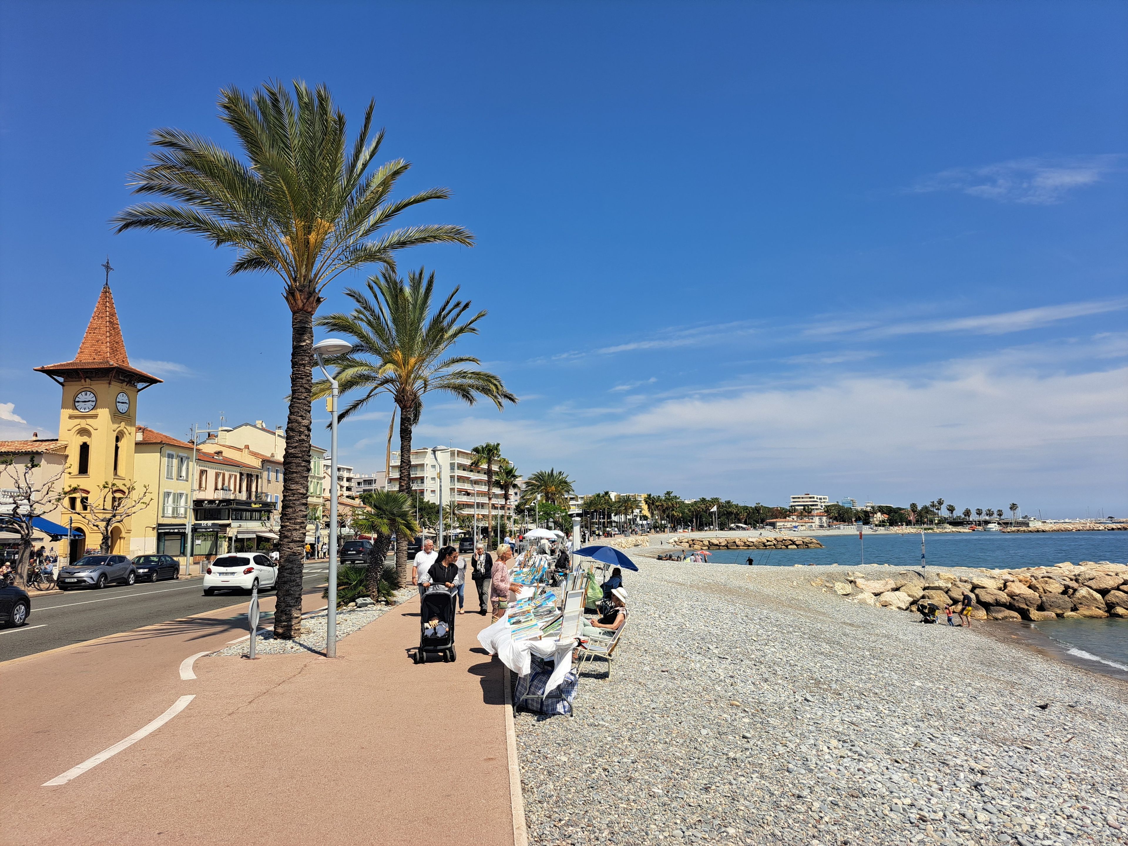 Un paseo marítimo con un campanario, palmeras y una playa de guijarros.