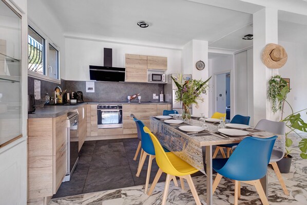 Una cocina moderna con electrodomésticos integrados, un comedor con mesa de mármol y sillas coloridas, y vistas al exterior.