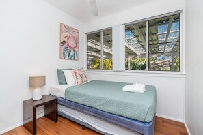 Ein Schlafzimmer mit einem Bett, einem Nachttisch, einer Lampe, einem Fenster mit Blick auf Grünflächen und einem Gemälde an der Wand.