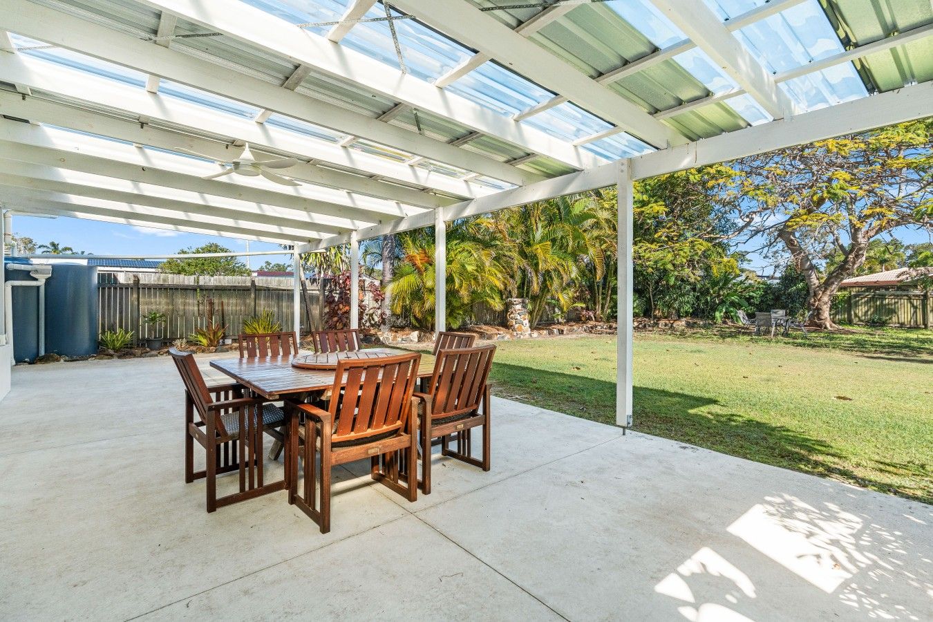 Une terrasse couverte, dotée d’une table et de chaises, entourée d’un jardin et d’une clôture en bois.
