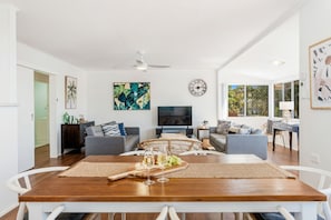 A spacious living room with a wooden dining table, grey sofas, a flat-screen TV, and a large window offering a view of greenery. - Ideal for Tradies. BBQ, WiFi, Pet friendly (Torquay)