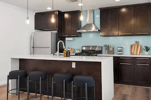 A modern kitchen with dark wood cabinets, a stainless steel range hood, a white refrigerator, and a bar area with stools. - Disney, Heated Pool, Game Rm, Fire Pit, King Bed- Getaway Retreat (Hacienda Heights)