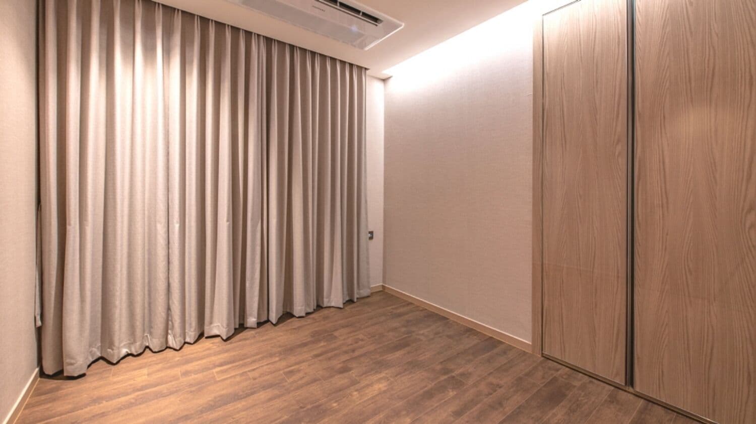 A room with a wooden floor, a large window with curtains, and a wardrobe.