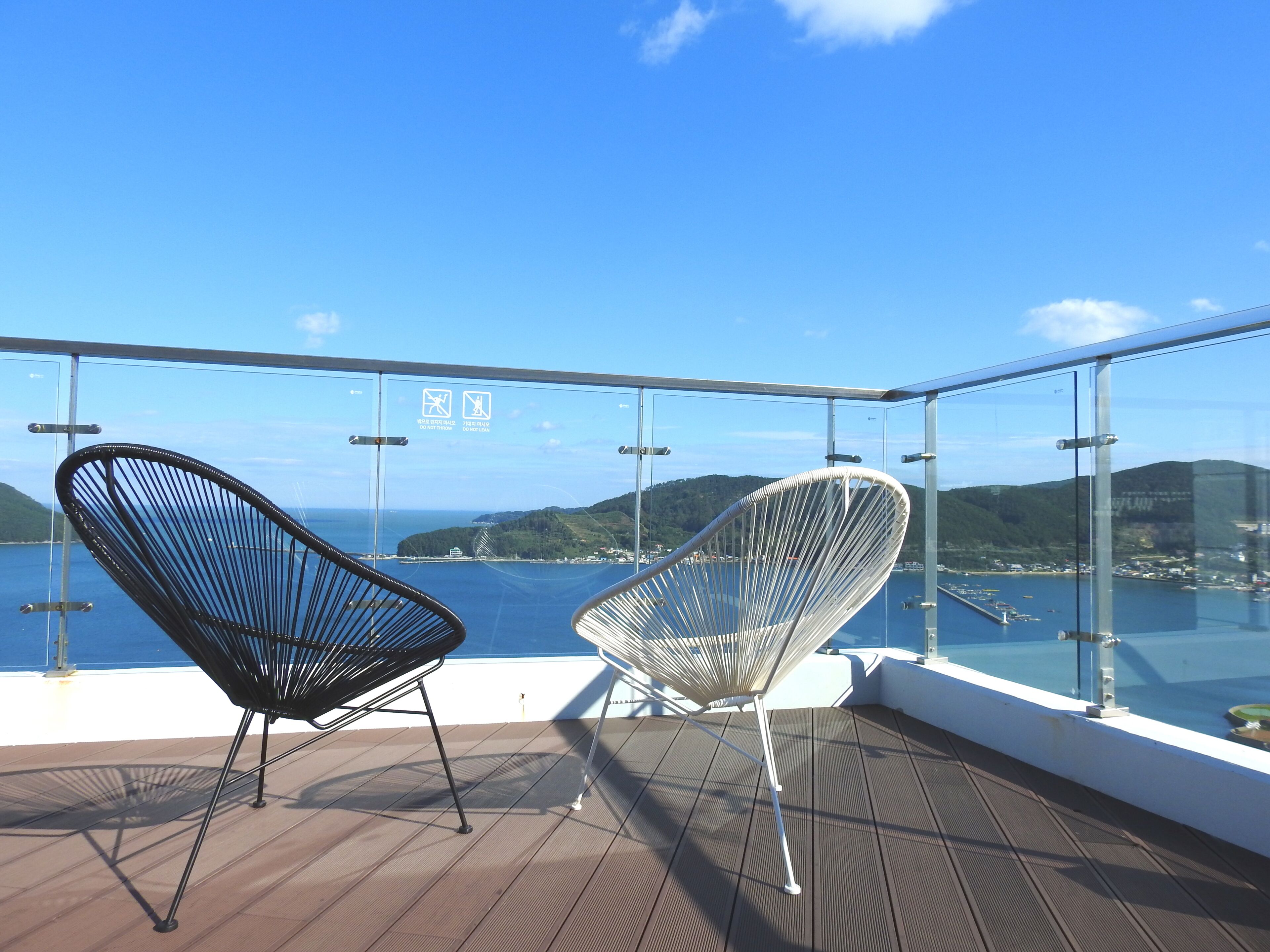 A balcony with two chairs, a glass railing, and a view of the sea and hills.