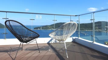 A balcony with two chairs, a glass railing, and a view of the sea and hills.