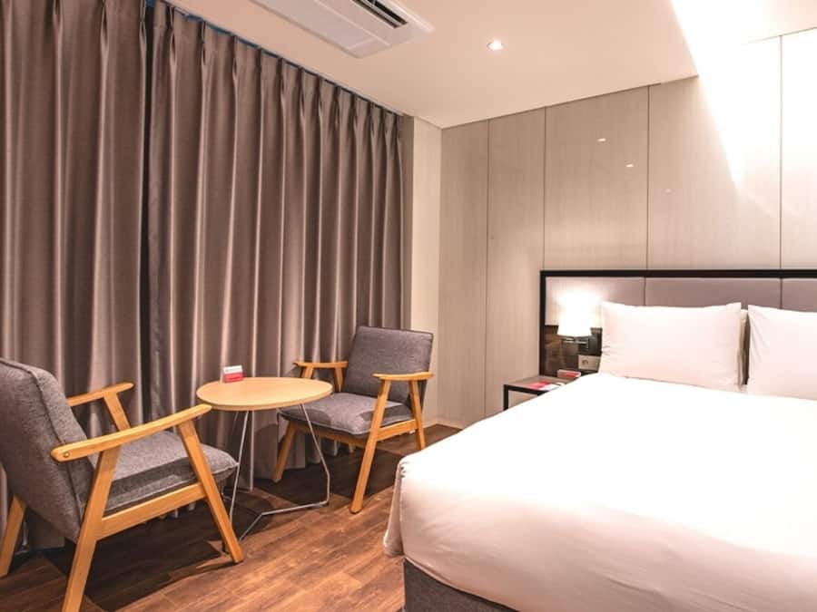 A hotel room with a bed, two armchairs, a small round table, and a nightstand with a lamp.