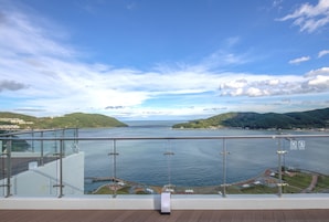 A panoramic view from a balcony overlooking a body of water, with a clear sky and distant hills.