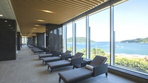 A sunlit lounge area with rows of lounge chairs facing a large window overlooking a coastal landscape.