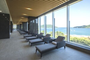 A sunlit lounge area with rows of lounge chairs facing a large window overlooking a coastal landscape.