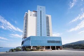 A modern hotel with a prominent glass facade, a central entrance, and a view of the sea. - Hotel The PORA Suitz (Geoje)
