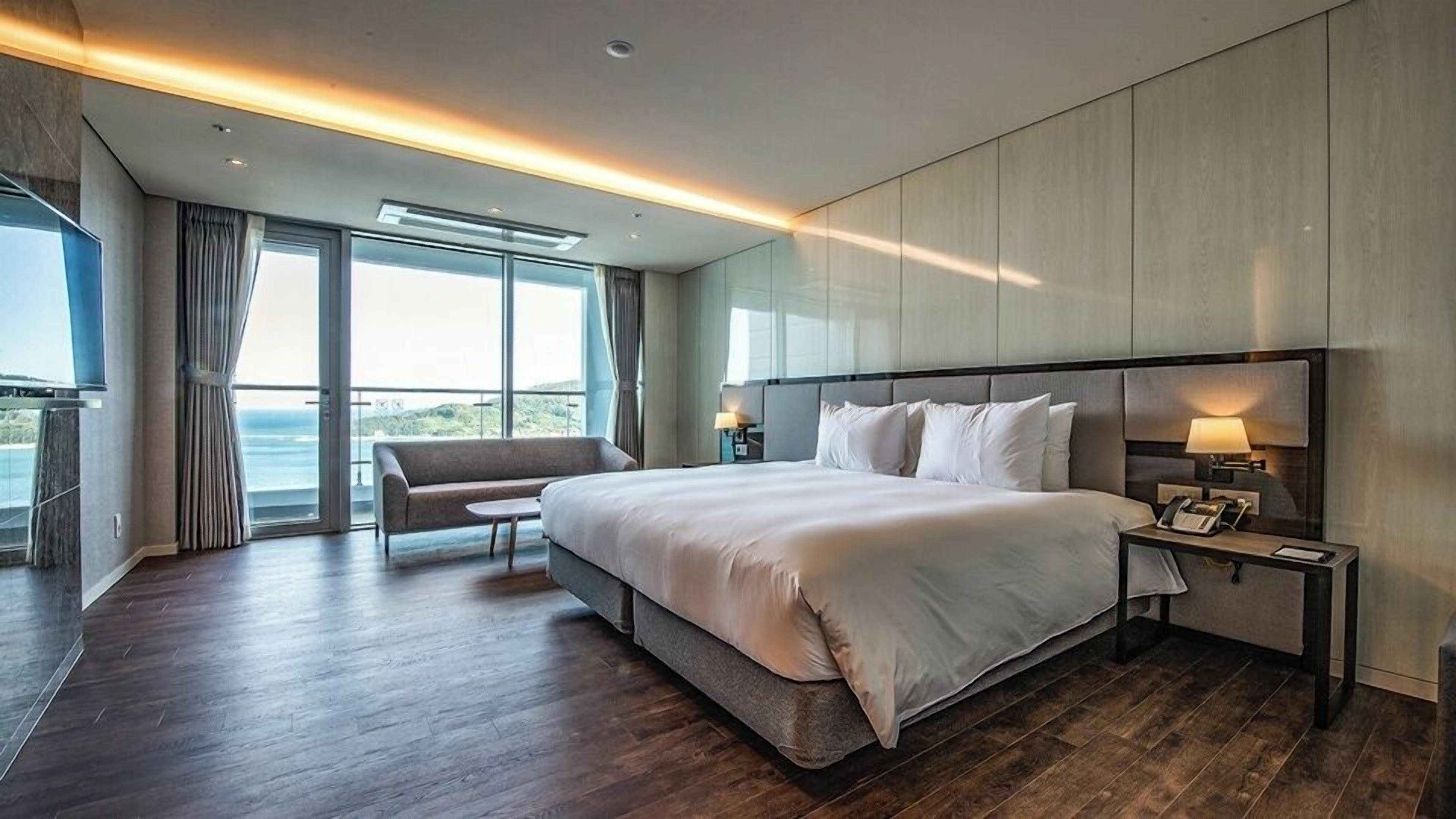 Una habitación de hotel moderna con una cama grande, un área de estar, una mesita de noche y vistas al mar.