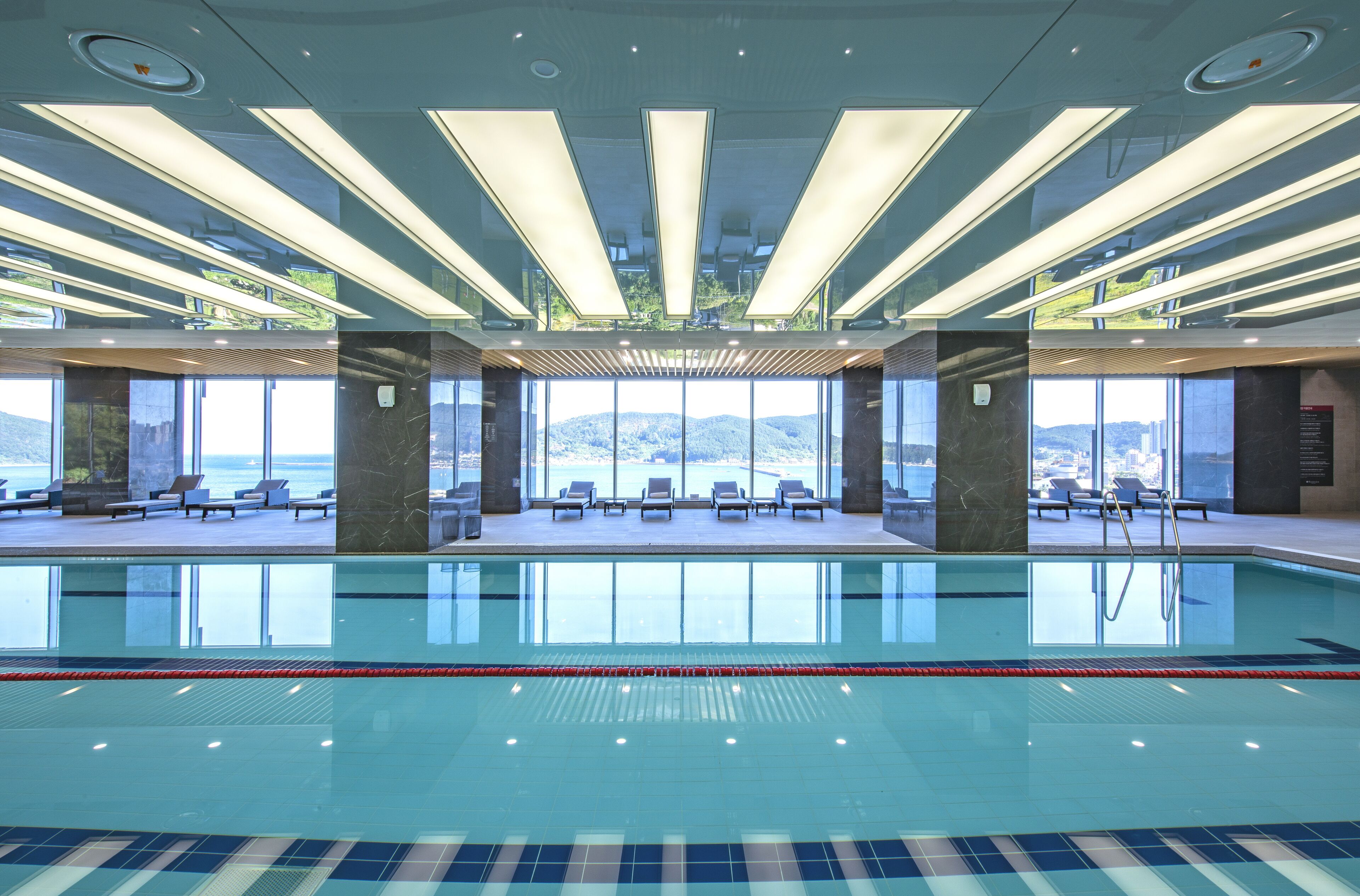A modern indoor swimming pool with clear water, sun loungers, and a view of the sea.