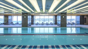 A modern indoor swimming pool with clear water, sun loungers, and a view of the sea.