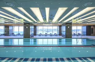 A modern indoor swimming pool with clear water, sun loungers, and a view of the sea.