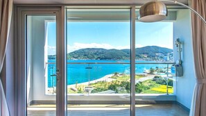 A room with a large sliding glass door leading to a balcony with a view of a coastal area and mountains.
