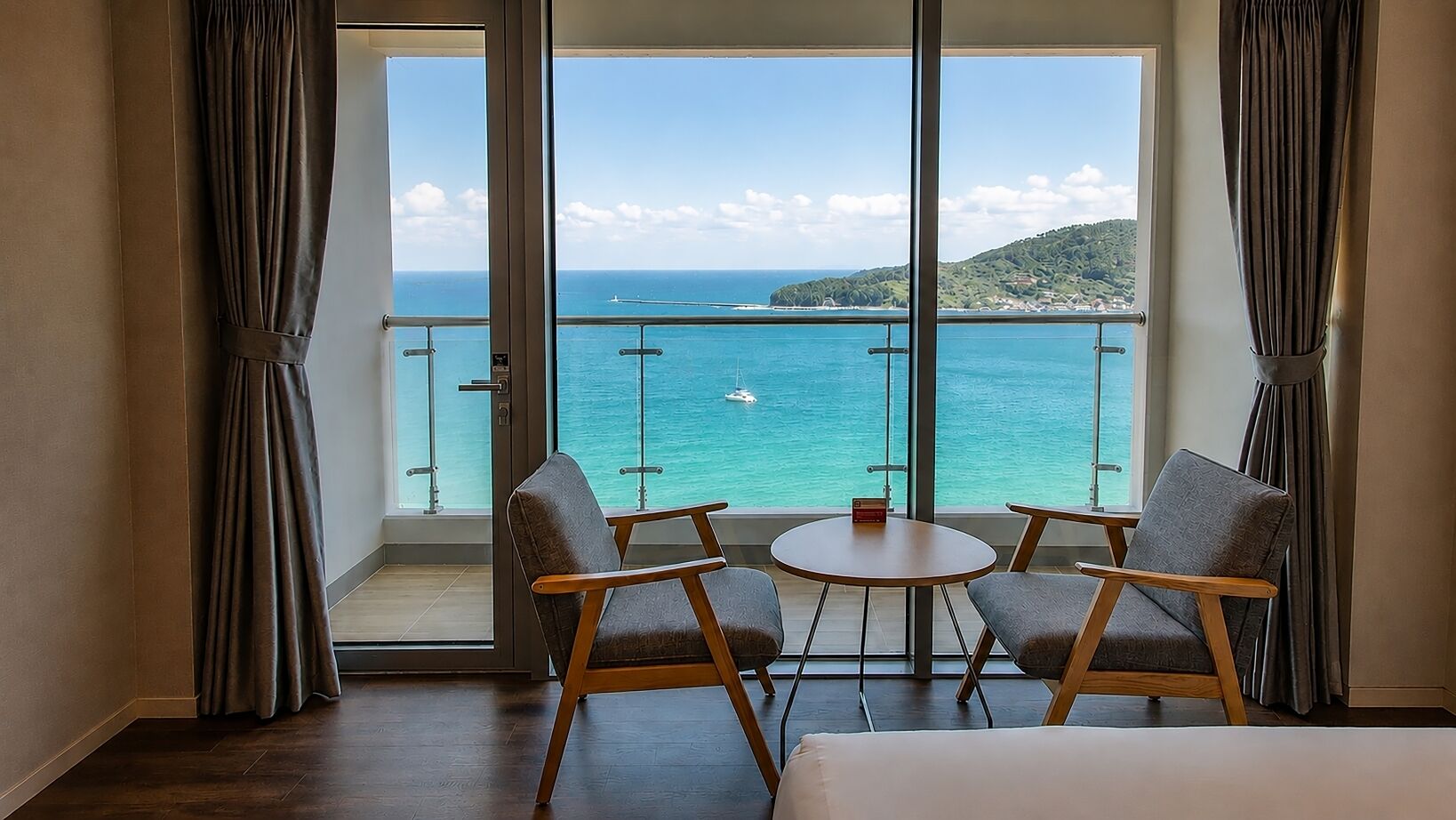 A room with a balcony overlooking a turquoise sea and a hilly landscape.