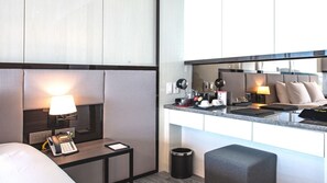 Hotelkamer met een bed, nachtkastje met een lamp, een telefoon, een klein tafeltje met een tablet, een kitchenette met een koffieapparaat en een prullenbak.