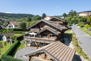Ein zweistöckiges Holzhaus mit einem Balkon, umgeben von einem gepflegten Garten und einer Straße.