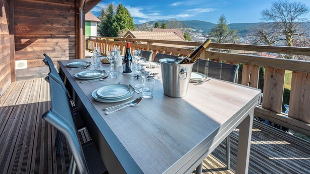 An outdoor dining area with a table set for four, a wine bottle, and a metal container, overlooking a scenic landscape. - Chalet 1907 (Gérardmer)