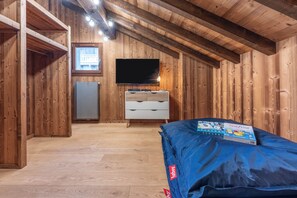 Ein Zimmer mit Holzwänden und schrägem Dach, einem an der Wand befestigten Flachbildfernseher, einer weißen Kommode mit Schubladen und einer blauen Matratze auf dem Boden.