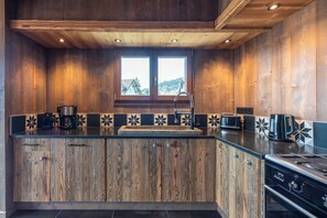 A kitchen with wooden cabinetry, a built-in sink, and a window with a view of a house and trees. - Chalet 1907 (Gérardmer)