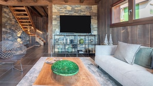 A modern living room with a stone fireplace, a flat-screen TV, a wooden staircase, and a sofa. - Chalet 1907 (Gérardmer)