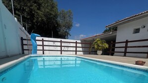 A swimming pool with a wooden fence, a palm tree, and a building with a tiled roof in the background. - Comfortable house with pool and 4 suites. Close to everything. Super quiet place. (Piranhas)