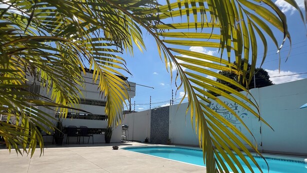 A swimming pool area with tiled flooring, palm trees, and a building with a blue-tiled wall in the background. - Comfortable house with pool and 4 suites. Close to everything. Super quiet place. (Piranhas)