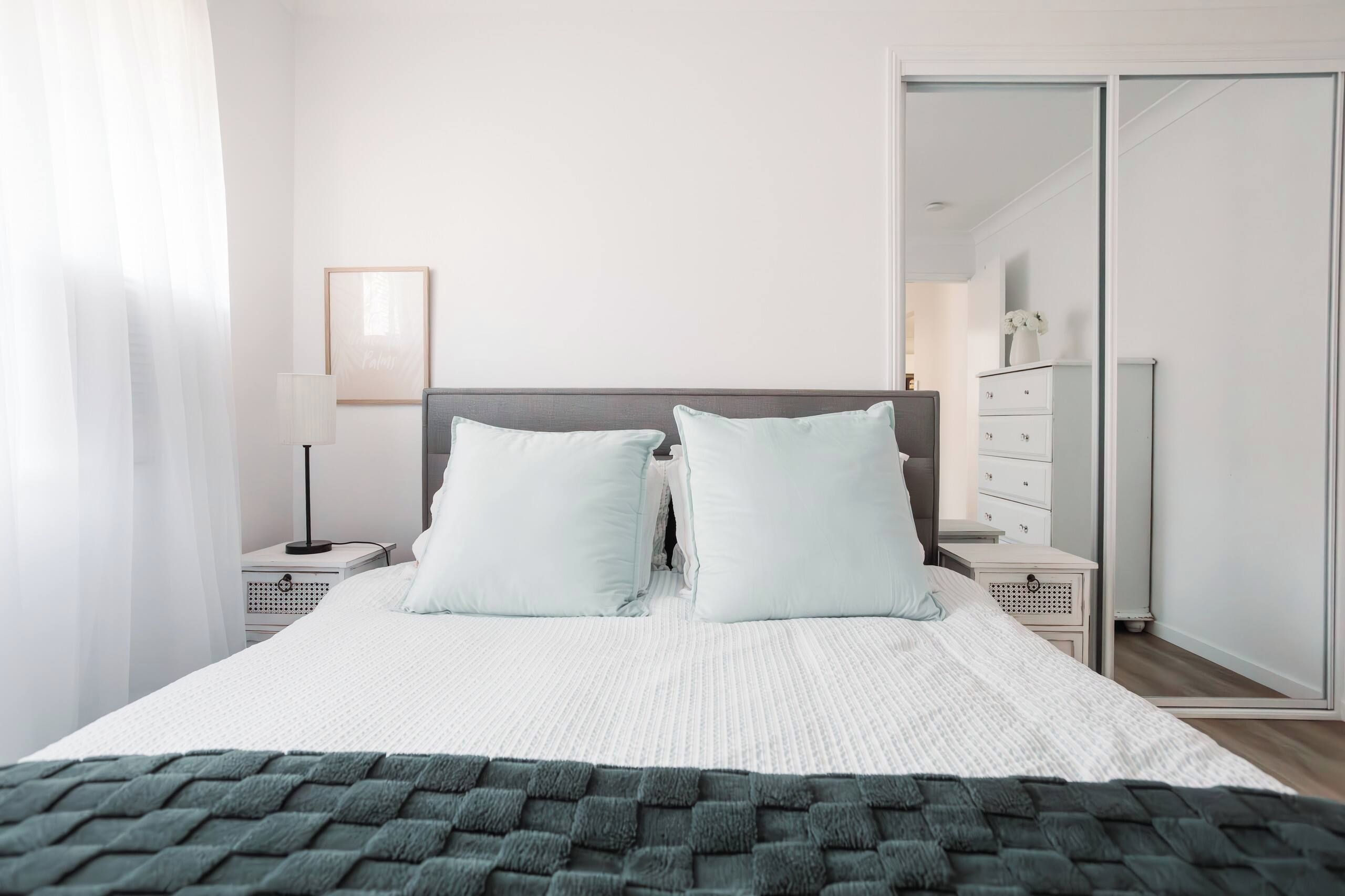 A neatly made bed with a white duvet and two pillows, a bedside table with a lamp, and a dresser with drawers.