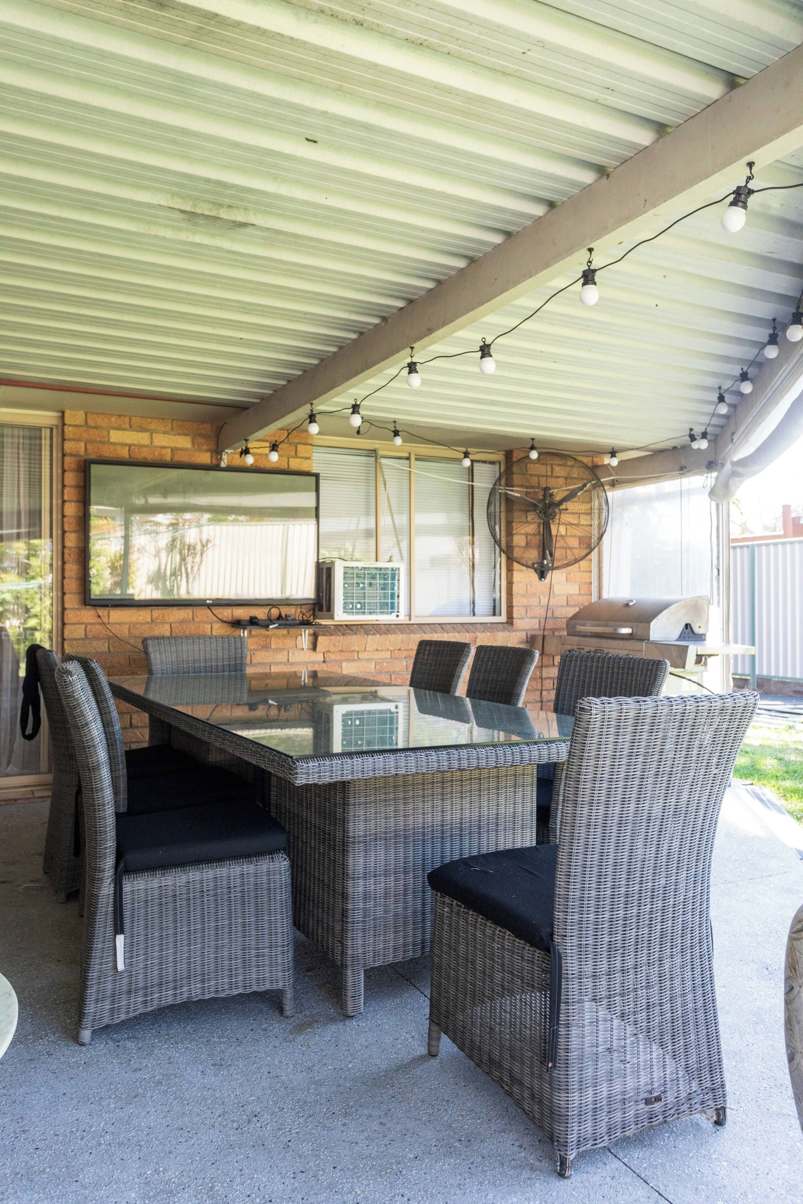 An outdoor patio with a glass-top table surrounded by wicker chairs, a ceiling fan, and string lights.
