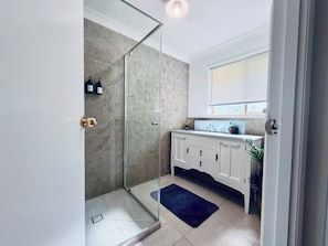 A modern bathroom with a glass shower enclosure, white cabinetry, and a window above the sink. - Serene Saltair - Large groups and family getaways (Redland Bay)