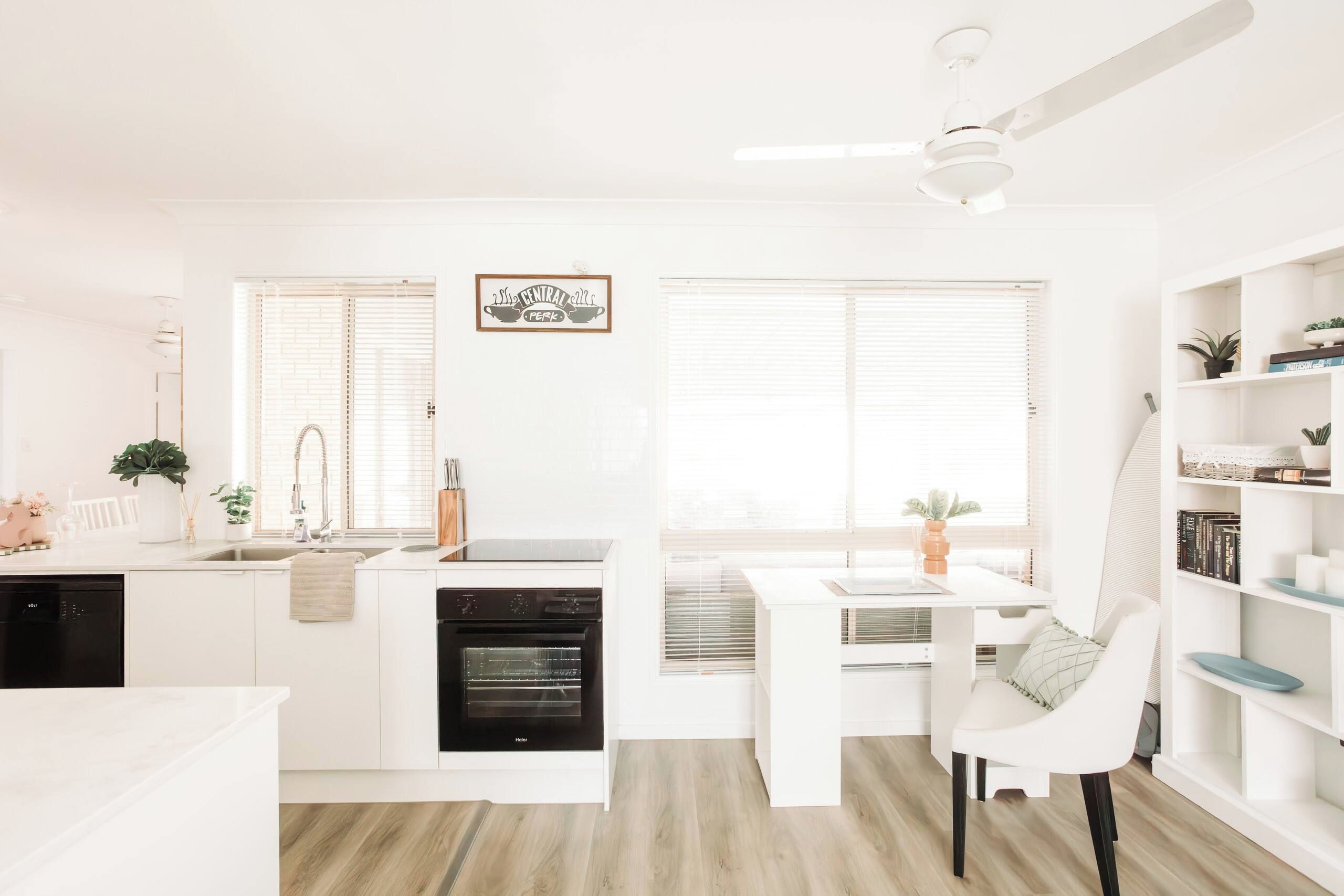 A modern kitchen with a central island, a dining area with a white table and chairs, and a bookshelf with various items.