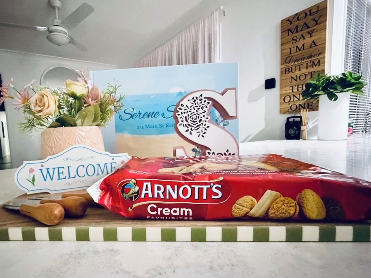 A kitchen counter with a welcome sign, a package of Arnott's Cream Favourites biscuits, a bouquet of flowers, and a decorative sign with a rose cut-out.