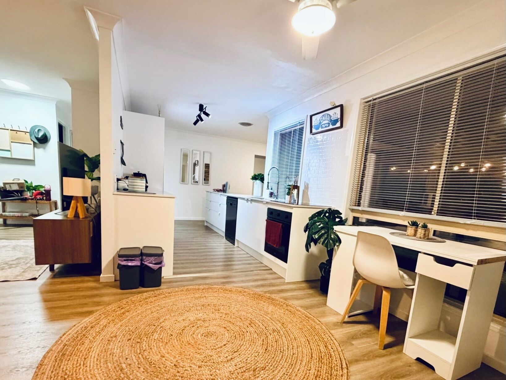 A modern living room with a large TV, a desk, and a comfortable chair. There is a kitchen area with a stove and a dining table.