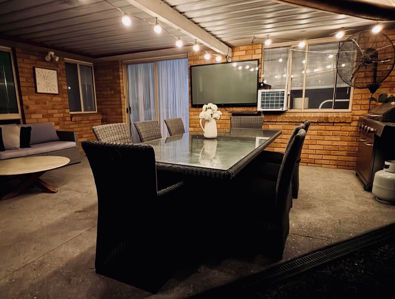 A cozy indoor patio with a glass-top table surrounded by chairs, a flat-screen TV mounted on the wall, and a grill area.