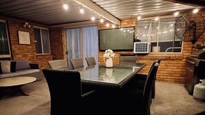 Un patio intérieur accueillant, doté d’une table à plateau en verre entourée de chaises, d’un téléviseur à écran plat fixé au mur et d’un espace barbecue.