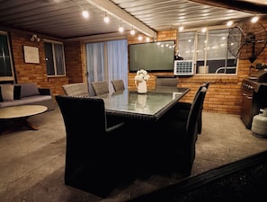 A cozy indoor patio with a glass-top table surrounded by chairs, a flat-screen TV mounted on the wall, and a grill area. - Serene Saltair - Large groups and family getaways (Redland Bay)