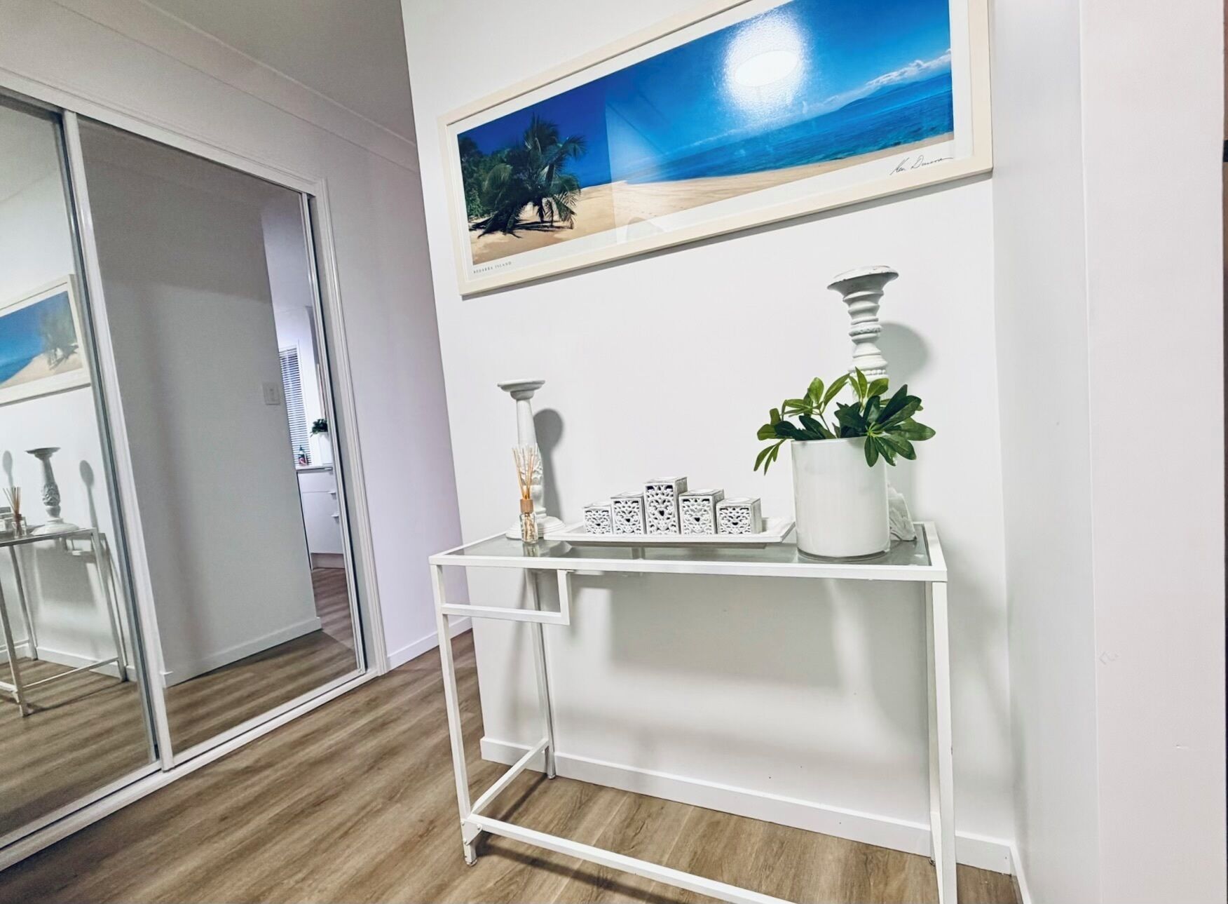 A room with a white console table, a framed picture of a beach, and a potted plant.