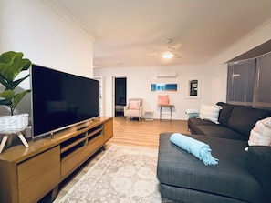 A living room with a flat-screen TV on a wooden stand, a dark sofa with a throw blanket, a patterned rug, and a ceiling fan. - Serene Saltair - Large groups and family getaways (Redland Bay)
