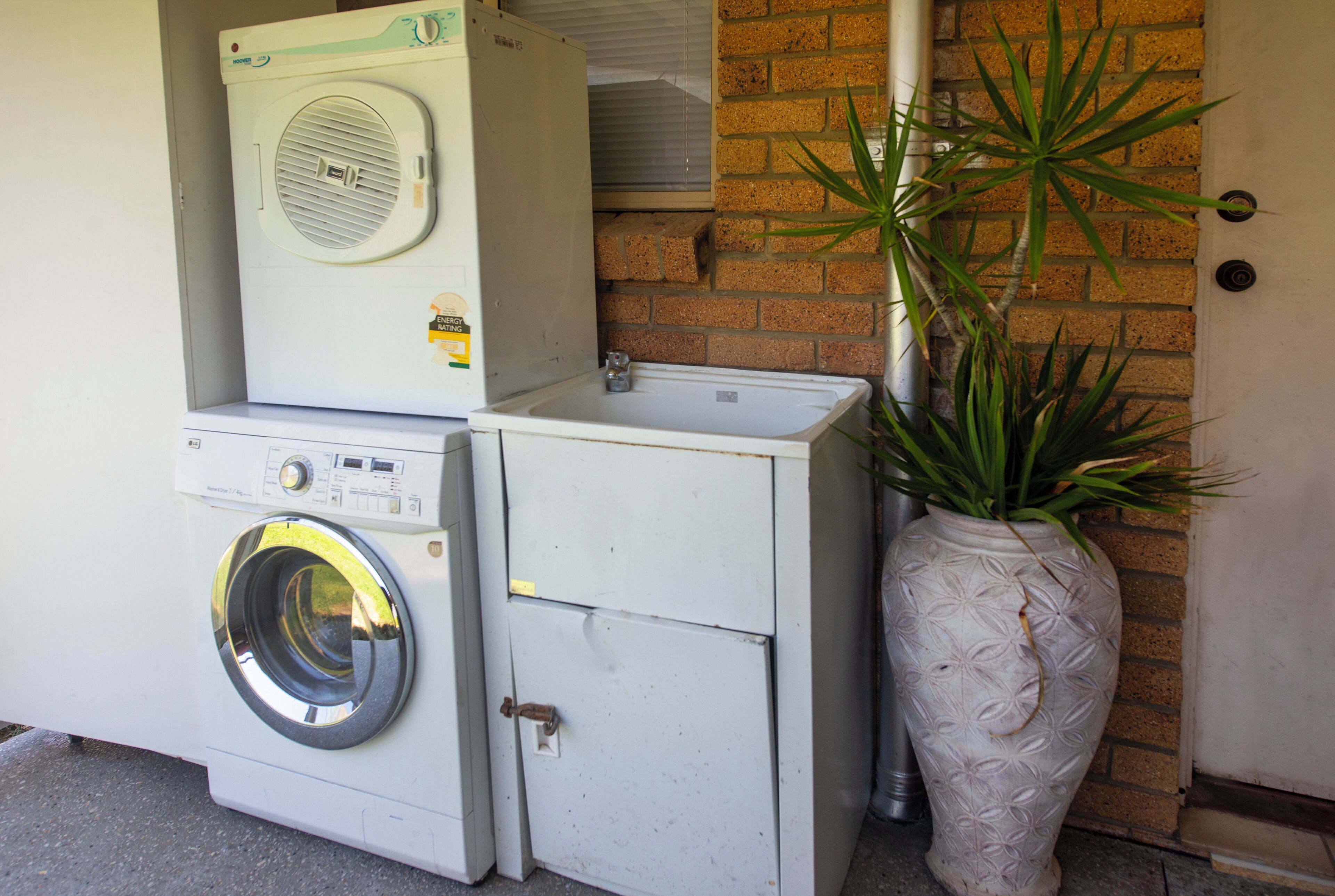 A front-loading washing machine and dryer stacked against a brick wall, with a potted plant beside them.