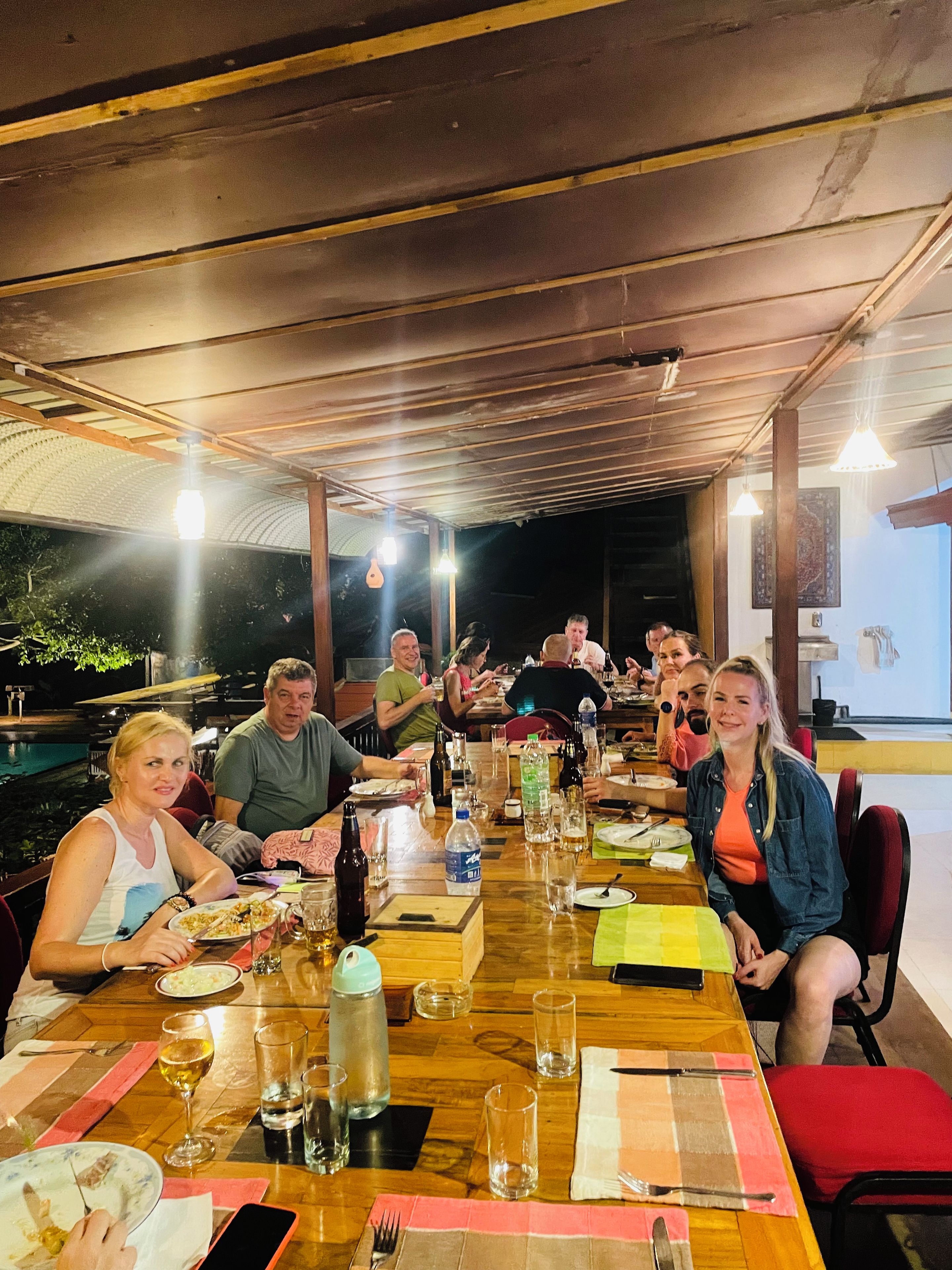 A group of people dining at an outdoor restaurant with a wooden table and chairs, surrounded by a covered area with a view of the outdoors.